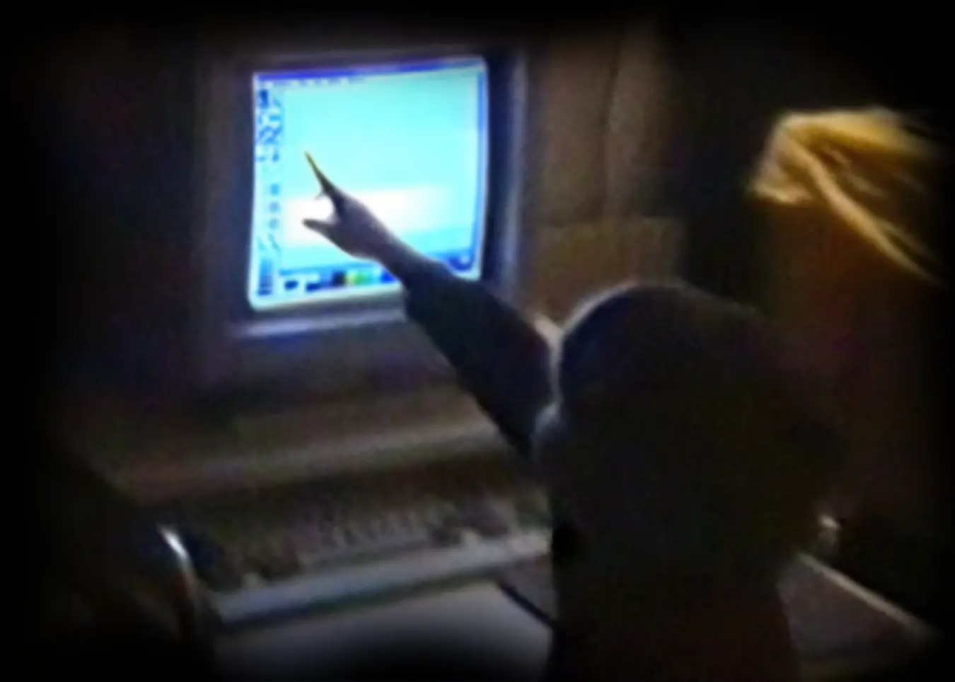 A dimly lit photo of Elias sitting at an old desktop computer, pointing at a bright blue screen running an early version of Microsoft Paint.