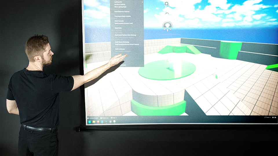 Elias leads a seminar, gesturing toward a large projector screen displaying a 3D virtual environment created in Unreal Engine.