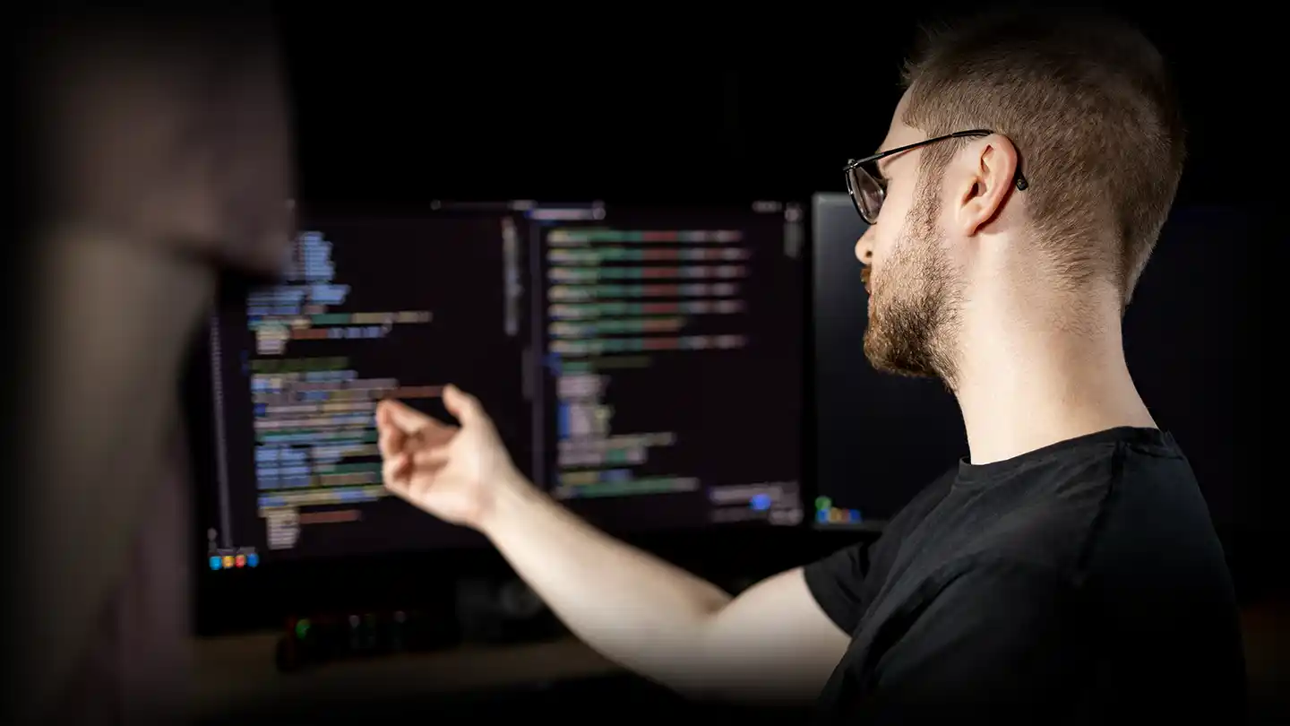 Elias sits at a desk in a dimly lit room, pointing at lines of code on a computer monitor while another person stands beside him.